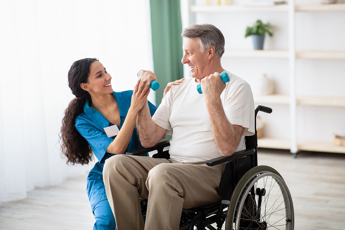 Doctor helping disabled elderly man in wheelchair to work out with dumbbells, to recover from injury at health centre